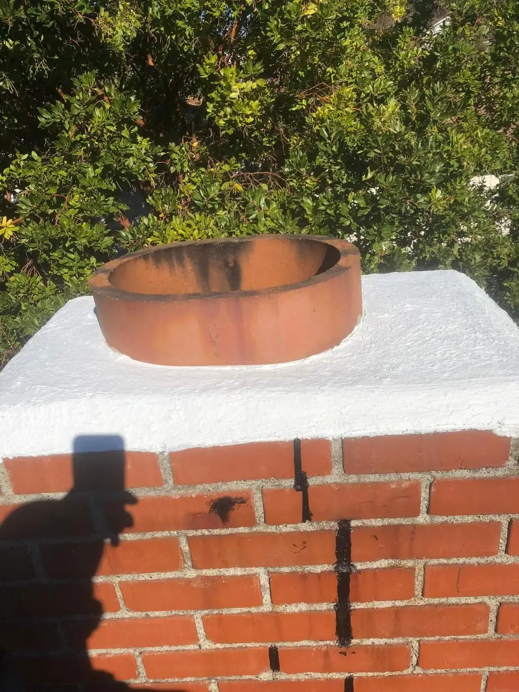 Una teja de conducto de barro en un remate blanco, sobre una chimenea de ladrillo rojo con manchas oscuras que bajan por el ladrillo.