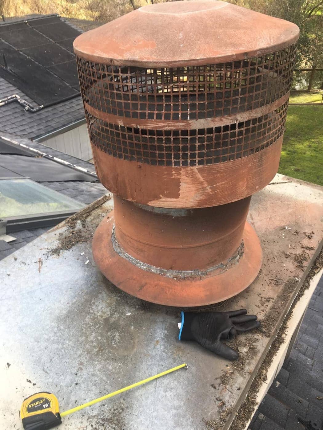 A round metal chimney cap with a mesh screen on a flat chimney top, with a work glove and a tape measure beside it.