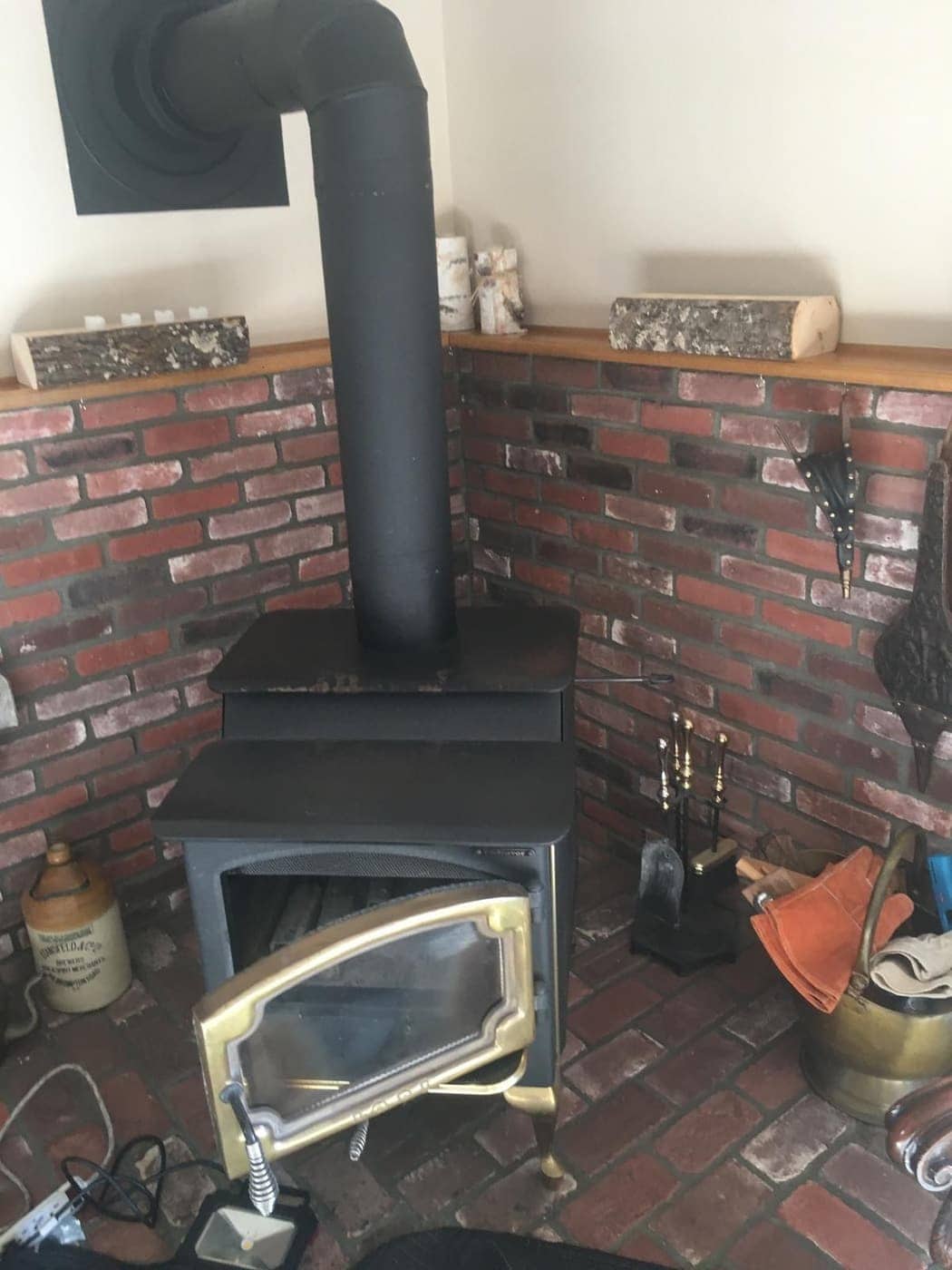 A wood stove with a black stovepipe, set in front of a brick wall.